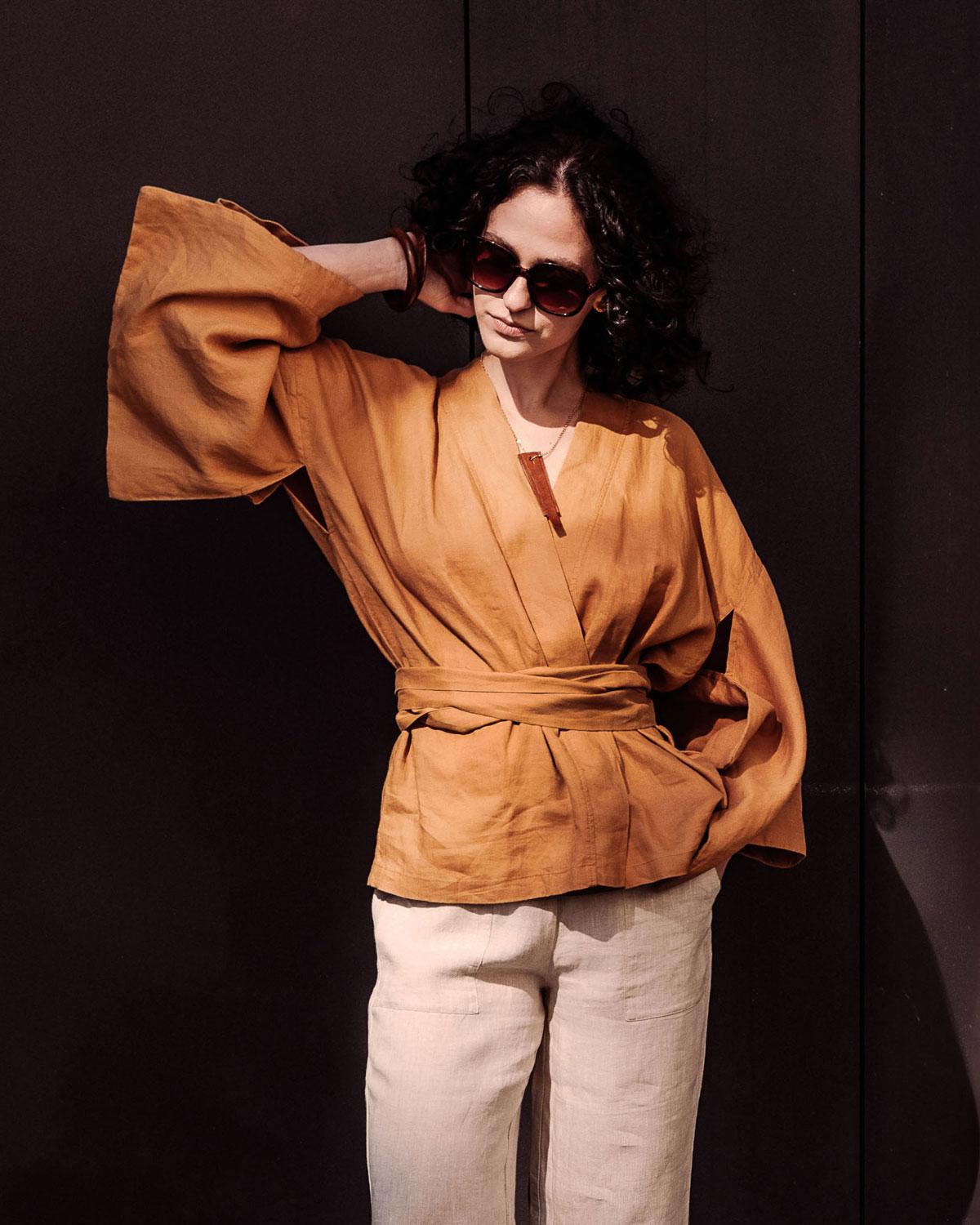 Kobieta w okularach przeciwsłonecznych i lnianym kimonie wiązanym w pasie w kolorze rudym (ochra) na ciemnym tle.